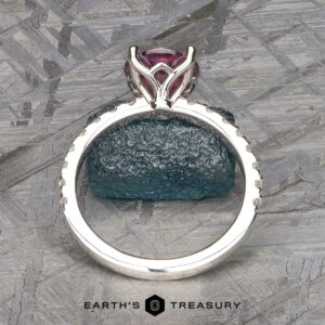 The Classic Pave "Blumia" Ring features a stunning large pink gemstone set in a silver band, which is adorned with small, sparkling diamonds. It is displayed upright against a textured background, with the text "Earth's Treasury" visible at the bottom center.
