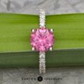 The Classic Pave "Blumia" Ring, featuring a central pink gemstone encircled by smaller clear stones along the silver band, is elegantly displayed resting on a green, leaf-like background. The text "Earth's Treasury" appears at the bottom of the image.
