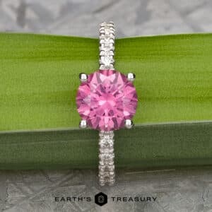 The Classic Pave "Blumia" Ring, featuring a central pink gemstone encircled by smaller clear stones along the silver band, is elegantly displayed resting on a green, leaf-like background. The text "Earth's Treasury" appears at the bottom of the image.