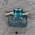 An exquisite ring called "The 'Waltz' Diamond-Accented Solitaire," featuring a large, square-cut teal gemstone, is displayed on a textured blue stone. The band showcases a twisted design with intricate detailing, and the logo "Earth's Treasury" is prominently visible at the bottom center of the image.