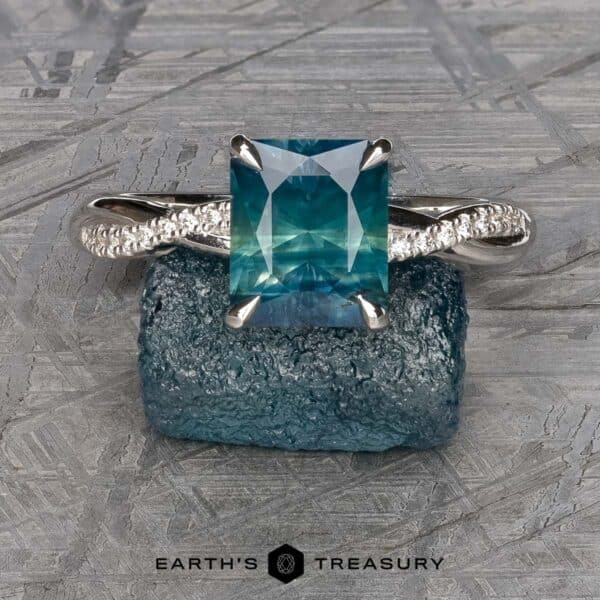 The Pave Waltz Ring in platinum with 2.70-carat Montana sapphire An exquisite ring called "The 'Waltz' Diamond-Accented Solitaire," featuring a large, square-cut teal gemstone, is displayed on a textured blue stone. The band showcases a twisted design with intricate detailing, and the logo "Earth's Treasury" is prominently visible at the bottom center of the image.