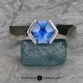 The "Ezra" Ring boasts a geometric band and a striking hexagonal blue gemstone at its center. It is showcased on a textured greenish-blue stone, with the "Earth's Treasury" logo appearing at the bottom of the image. The background features a metallic, etched pattern.