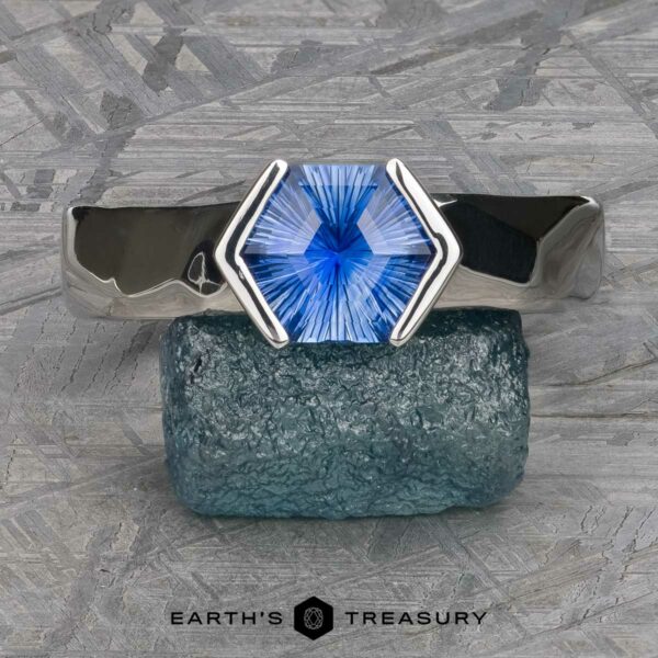 The Ezra Ring in platinum with 1.53-Carat Ceylon Sapphire The "Ezra" Ring boasts a geometric band and a striking hexagonal blue gemstone at its center. It is showcased on a textured greenish-blue stone, with the "Earth's Treasury" logo appearing at the bottom of the image. The background features a metallic, etched pattern.