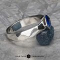 The "Ezra" Ring boasts a wavy silver band, adorned with a blue gemstone set diagonally at its center. The ring is elegantly displayed on a textured dark-grey stone, against a background that features a detailed, patterned surface with the "Earth's Treasury" logo and text at the bottom.