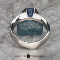 A silver ring named "Ezra," adorned with a blue gemstone, is set against a textured gray and blue stone backdrop. The ring features a unique geometric design, showcasing the blue gemstone in a modern, angled setting. The image includes the text "Earth's Treasury" at the bottom.
