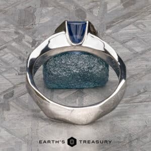 A silver ring named "Ezra," adorned with a blue gemstone, is set against a textured gray and blue stone backdrop. The ring features a unique geometric design, showcasing the blue gemstone in a modern, angled setting. The image includes the text "Earth's Treasury" at the bottom.