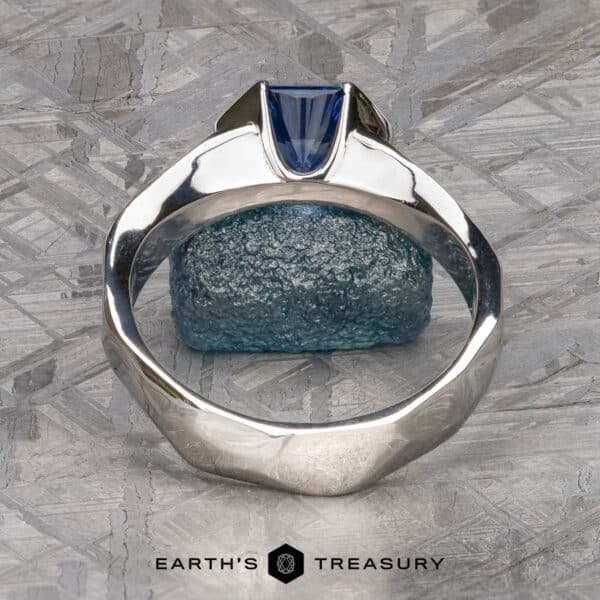 The Ezra Ring in platinum with 1.53-Carat Ceylon Sapphire A silver ring named "Ezra," adorned with a blue gemstone, is set against a textured gray and blue stone backdrop. The ring features a unique geometric design, showcasing the blue gemstone in a modern, angled setting. The image includes the text "Earth's Treasury" at the bottom.