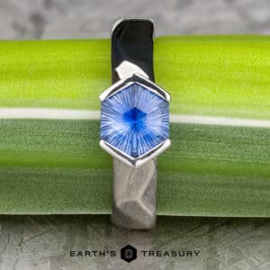 A close-up of "The Ezra Ring" showcasing its silver band and blue hexagon-shaped gemstone with intricate facets. The ring is placed on a green background, with the text "EARTH'S TREASURY" visible at the bottom of the image.