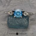The “Minerva” Three-Stone Ring, a silver piece showcasing a round blue gemstone flanked by two smaller clear gemstones, is displayed on a textured blue-green rock. The background consists of a gray, patterned surface. The text "EARTH'S TREASURY" appears below the image.