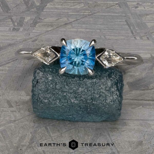 The "Minerva" Ring in 14k white gold with 0.98-carat Montana sapphire The “Minerva” Three-Stone Ring, a silver piece showcasing a round blue gemstone flanked by two smaller clear gemstones, is displayed on a textured blue-green rock. The background consists of a gray, patterned surface. The text "EARTH'S TREASURY" appears below the image.
