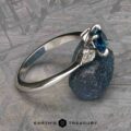 The “Minerva” Three-Stone Ring features a stunning silver band adorned with a central blue gemstone and detailed with white square gemstones. It is beautifully displayed on a textured blue stone. The background is a light-colored, patterned surface, complete with the brand name "Earth's Treasury" printed at the bottom.