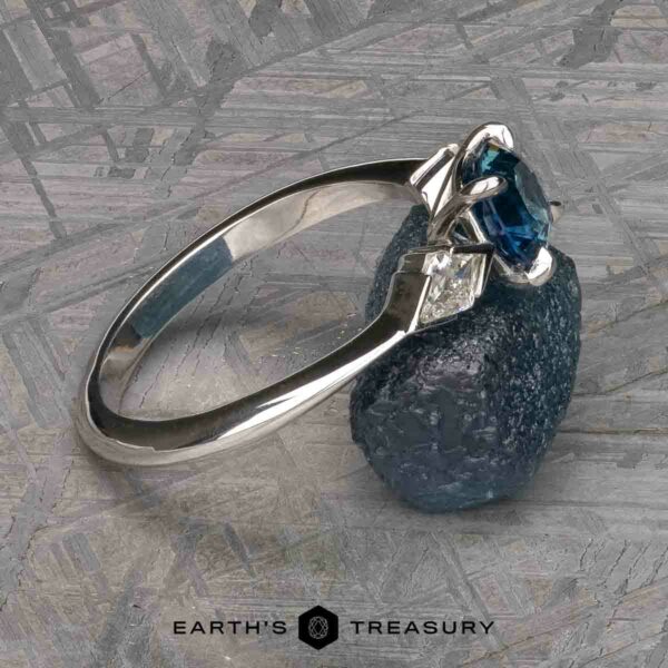 The "Minerva" Ring in 14k white gold with 0.98-carat Montana sapphire The “Minerva” Three-Stone Ring features a stunning silver band adorned with a central blue gemstone and detailed with white square gemstones. It is beautifully displayed on a textured blue stone. The background is a light-colored, patterned surface, complete with the brand name "Earth's Treasury" printed at the bottom.