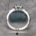 The "Minerva" Three-Stone Ring, showcasing a central blue gemstone secured in a four-prong setting, is displayed upright. It rests atop a textured blue stone, with an intricate pattern serving as the backdrop. Below the ring, the text "Earth's Treasury" is prominently visible.