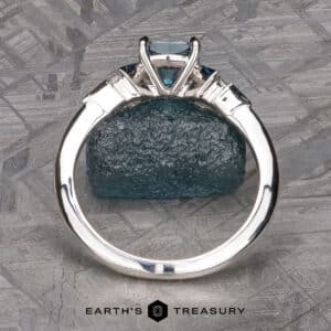 The "Minerva" Three-Stone Ring, showcasing a central blue gemstone secured in a four-prong setting, is displayed upright. It rests atop a textured blue stone, with an intricate pattern serving as the backdrop. Below the ring, the text "Earth's Treasury" is prominently visible.