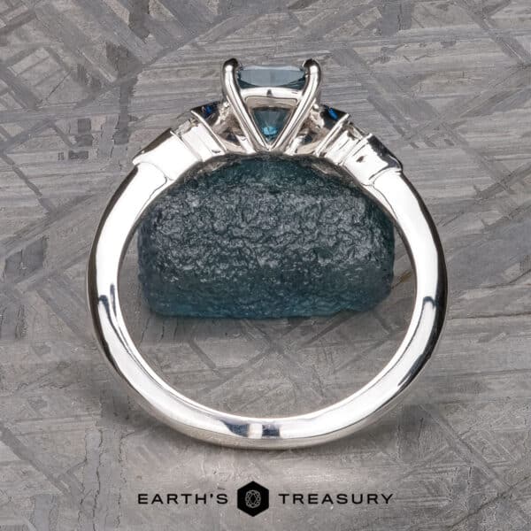 The "Minerva" Ring in 14k white gold with 0.98-carat Montana sapphire The "Minerva" Three-Stone Ring, showcasing a central blue gemstone secured in a four-prong setting, is displayed upright. It rests atop a textured blue stone, with an intricate pattern serving as the backdrop. Below the ring, the text "Earth's Treasury" is prominently visible.