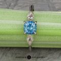 The “Minerva” Three-Stone Ring showcases a blue round-cut Montana sapphire as its centerpiece, flanked by two diamond accent stones on either side. The ring is displayed on a green and white striped background with the "Earth's Treasury" logo positioned at the bottom of the image.