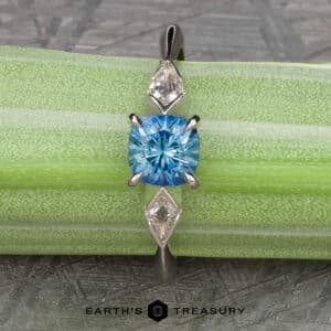 The “Minerva” Three-Stone Ring showcases a blue round-cut Montana sapphire as its centerpiece, flanked by two diamond accent stones on either side. The ring is displayed on a green and white striped background with the "Earth's Treasury" logo positioned at the bottom of the image.