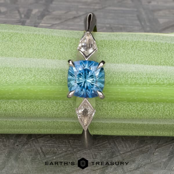 The "Minerva" Ring in 14k white gold with 0.98-carat Montana sapphire The “Minerva” Three-Stone Ring showcases a blue round-cut Montana sapphire as its centerpiece, flanked by two diamond accent stones on either side. The ring is displayed on a green and white striped background with the "Earth's Treasury" logo positioned at the bottom of the image.