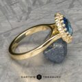 A close-up shot of The Prong-Set "Valeria" Halo Ring showcases a large blue gemstone encircled by small diamonds. Resting on a metallic surface and supported by a small rock, the ring exudes elegance. At the bottom of the image, the logo and text "Earth's Treasury" are displayed.