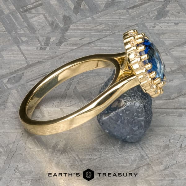 The Prong-Set Valeria ring in 18k yellow gold with 2.96-carat Madagascar sapphire A close-up shot of The Prong-Set "Valeria" Halo Ring showcases a large blue gemstone encircled by small diamonds. Resting on a metallic surface and supported by a small rock, the ring exudes elegance. At the bottom of the image, the logo and text "Earth's Treasury" are displayed.