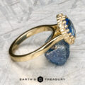 A close-up shot of The Prong-Set "Valeria" Halo Ring showcases a large blue gemstone encircled by small diamonds. Resting on a metallic surface and supported by a small rock, the ring exudes elegance. At the bottom of the image, the logo and text "Earth's Treasury" are displayed.