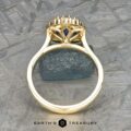 The Prong-Set "Valeria" Halo Ring features a meticulous, ornate design showcasing a striking blue gemstone at its core, captured from a side perspective. The ring rests on a textured gray surface with an additional small blue stone as an accent. The text "Earth's Treasury" is displayed at the bottom.