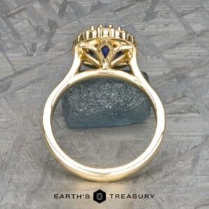 The Prong-Set "Valeria" Halo Ring features a meticulous, ornate design showcasing a striking blue gemstone at its core, captured from a side perspective. The ring rests on a textured gray surface with an additional small blue stone as an accent. The text "Earth's Treasury" is displayed at the bottom.