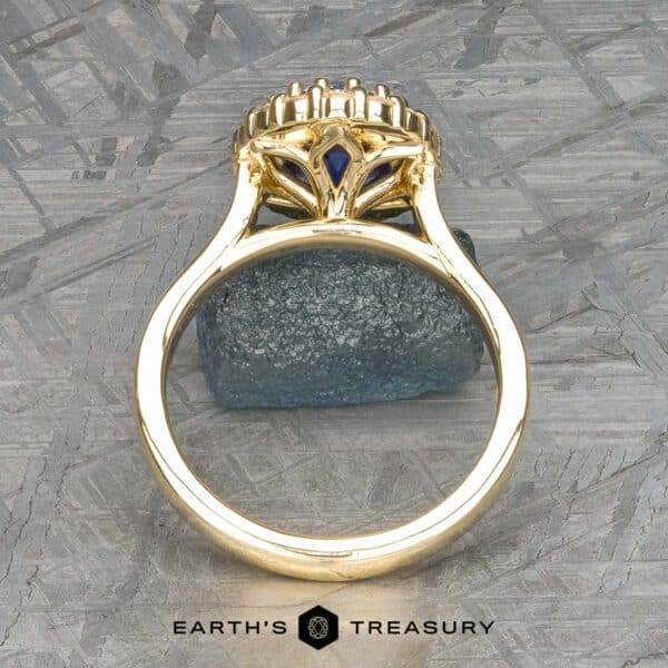 The Prong-Set Valeria ring in 18k yellow gold with 2.96-carat Madagascar sapphire The Prong-Set "Valeria" Halo Ring features a meticulous, ornate design showcasing a striking blue gemstone at its core, captured from a side perspective. The ring rests on a textured gray surface with an additional small blue stone as an accent. The text "Earth's Treasury" is displayed at the bottom.