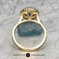 The Prong-Set "Valeria" Halo Ring features a meticulous, ornate design showcasing a striking blue gemstone at its core, captured from a side perspective. The ring rests on a textured gray surface with an additional small blue stone as an accent. The text "Earth's Treasury" is displayed at the bottom.