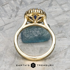 The Prong-Set "Valeria" Halo Ring features a meticulous, ornate design showcasing a striking blue gemstone at its core, captured from a side perspective. The ring rests on a textured gray surface with an additional small blue stone as an accent. The text "Earth's Treasury" is displayed at the bottom.