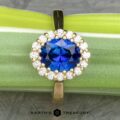 The Prong-Set "Valeria" Halo Ring is showcased against a green and silver background. The central vibrant blue gemstone is surrounded by a halo of small, round, white diamonds, all set in a gold band. The text "EARTH'S TREASURY" is displayed at the bottom.