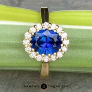 The Prong-Set "Valeria" Halo Ring is showcased against a green and silver background. The central vibrant blue gemstone is surrounded by a halo of small, round, white diamonds, all set in a gold band. The text "EARTH'S TREASURY" is displayed at the bottom.