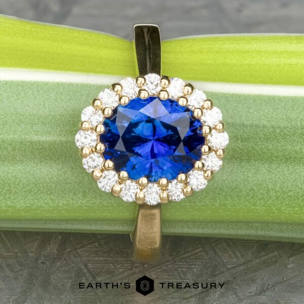 The Prong-Set Valeria ring in 18k yellow gold with 2.96-carat Madagascar sapphire The Prong-Set "Valeria" Halo Ring is showcased against a green and silver background. The central vibrant blue gemstone is surrounded by a halo of small, round, white diamonds, all set in a gold band. The text "EARTH'S TREASURY" is displayed at the bottom.