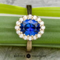 The Prong-Set "Valeria" Halo Ring, featuring a large blue sapphire center stone encircled by small white diamonds, is displayed on a textured stone surface. The ring's band is crafted from polished metal, with the text "EARTH'S TREASURY" visible at the bottom of the image.