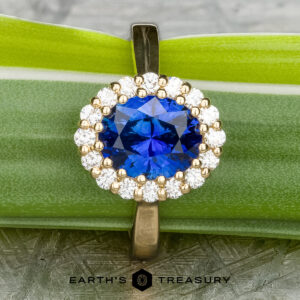 The Prong-Set "Valeria" Halo Ring, featuring a large blue sapphire center stone encircled by small white diamonds, is displayed on a textured stone surface. The ring's band is crafted from polished metal, with the text "EARTH'S TREASURY" visible at the bottom of the image.