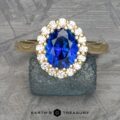 The Prong-Set "Valeria" Halo Ring, featuring a large blue sapphire center stone encircled by small white diamonds, is displayed on a textured stone surface. The ring's band is crafted from polished metal, with the text "EARTH'S TREASURY" visible at the bottom of the image.