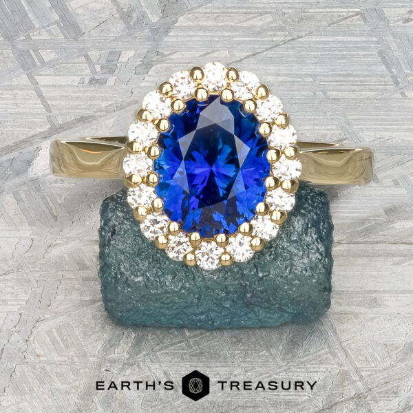 The Prong-Set Valeria ring in 18k yellow gold with 2.96-carat Madagascar sapphire The Prong-Set "Valeria" Halo Ring, featuring a large blue sapphire center stone encircled by small white diamonds, is displayed on a textured stone surface. The ring's band is crafted from polished metal, with the text "EARTH'S TREASURY" visible at the bottom of the image.