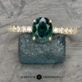 The Classic Pave "Eugenie" Hidden Halo Ring with a large, oval-cut teal gemstone is displayed on a textured stone. The ring features a band encrusted with small diamonds. The background showcases a patterned, metallic surface, while the text "EARTH'S TREASURY" appears at the bottom of the image.