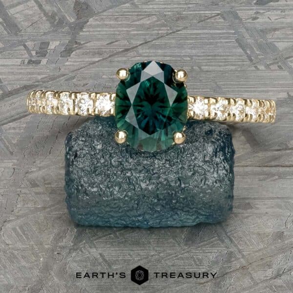 The Classic Pave Eugenie Ring in 14k yellow gold with 1.65-carat Australian sapphire The Classic Pave "Eugenie" Hidden Halo Ring with a large, oval-cut teal gemstone is displayed on a textured stone. The ring features a band encrusted with small diamonds. The background showcases a patterned, metallic surface, while the text "EARTH'S TREASURY" appears at the bottom of the image.