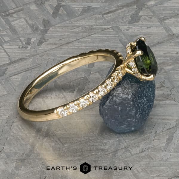 The Classic Pave Eugenie Ring in 14k yellow gold with 1.65-carat Australian sapphire The Classic Pave "Eugenie" Hidden Halo Ring, featuring a dark green marquise-cut gemstone set in gold, is elegantly placed on a small blue-gray stone. The ring's band is embellished with numerous small white diamonds. The text "EARTH'S TREASURY" is displayed at the bottom of the image.