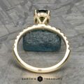 A gold Classic Pave "Eugenie" Hidden Halo Ring set with a dark central gemstone and smaller clear stones is situated on a textured surface. In the background, a chunk of raw blue-green mineral is visible. At the bottom, the text reads: "EARTH'S TREASURY.