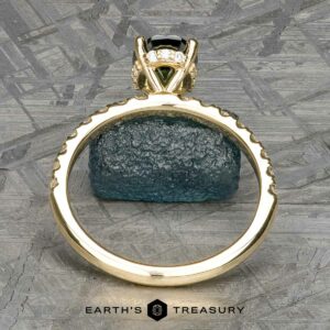 A gold Classic Pave "Eugenie" Hidden Halo Ring set with a dark central gemstone and smaller clear stones is situated on a textured surface. In the background, a chunk of raw blue-green mineral is visible. At the bottom, the text reads: "EARTH'S TREASURY.