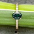 A gold ring, named The Classic Pave "Eugenie" Hidden Halo Ring, features a round, dark green gemstone set at its center. The band is adorned with clear diamonds and the ring is elegantly displayed on a light green plant stem. The image includes the logo and text "EARTH'S TREASURY" at the bottom.