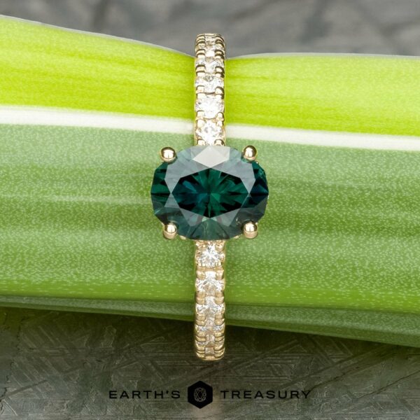 The Classic Pave Eugenie Ring in 14k yellow gold with 1.65-carat Australian sapphire A gold ring, named The Classic Pave "Eugenie" Hidden Halo Ring, features a round, dark green gemstone set at its center. The band is adorned with clear diamonds and the ring is elegantly displayed on a light green plant stem. The image includes the logo and text "EARTH'S TREASURY" at the bottom.