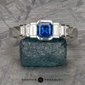 The "Apollo" Ring is a stunning silver ring featuring a central emerald-cut blue gemstone, complemented by smaller rectangular clear stones on either side. It is elegantly showcased on a textured blue stone with a silver patterned background. The text "Earth's Treasury" is prominently displayed at the bottom center.