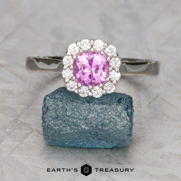 The Prong "Valeria" ring in 18K white gold with 1.14-carat Madagascar sapphire The Rosenthal, Aaron ring features a round pink gemstone prong-set, surrounded by small diamonds in silver, displayed on rough blue crystal. Set against a light gray background with "EARTH'S TREASURY" text below.