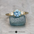 The Cathedral "Merope" ring in 18k yellow gold, set with a 1.24-carat blue oval gemstone and two clear side stones, is displayed on a rough blue crystal with the "Earth's Treasury" logo at the bottom.