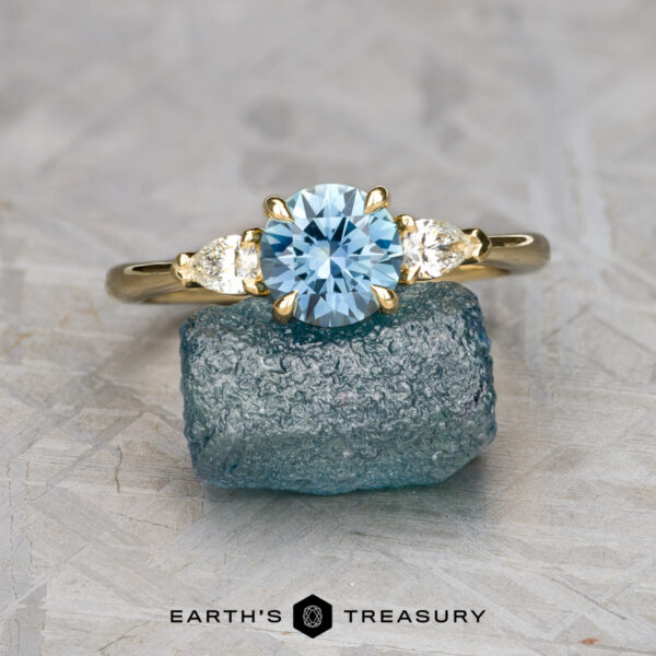 The Cathedral "Merope" ring in 18K yellow gold with 1.24-carat Montana sapphire The Cathedral "Merope" ring in 18k yellow gold, set with a 1.24-carat blue oval gemstone and two clear side stones, is displayed on a rough blue crystal with the "Earth's Treasury" logo at the bottom.