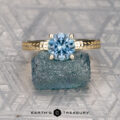 The "Kerria" ring in 14k yellow gold with a 1.40-carat Montana sapphire rests on a rough blue stone atop a patterned metallic surface, with the "Earth's Treasury" logo below.