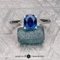 The "Vesta" ring in platinum, set with a 2.44-carat Montana sapphire, is shown on a rough blue stone against a geometric metallic background featuring the "EARTH'S TREASURY" logo at the bottom.